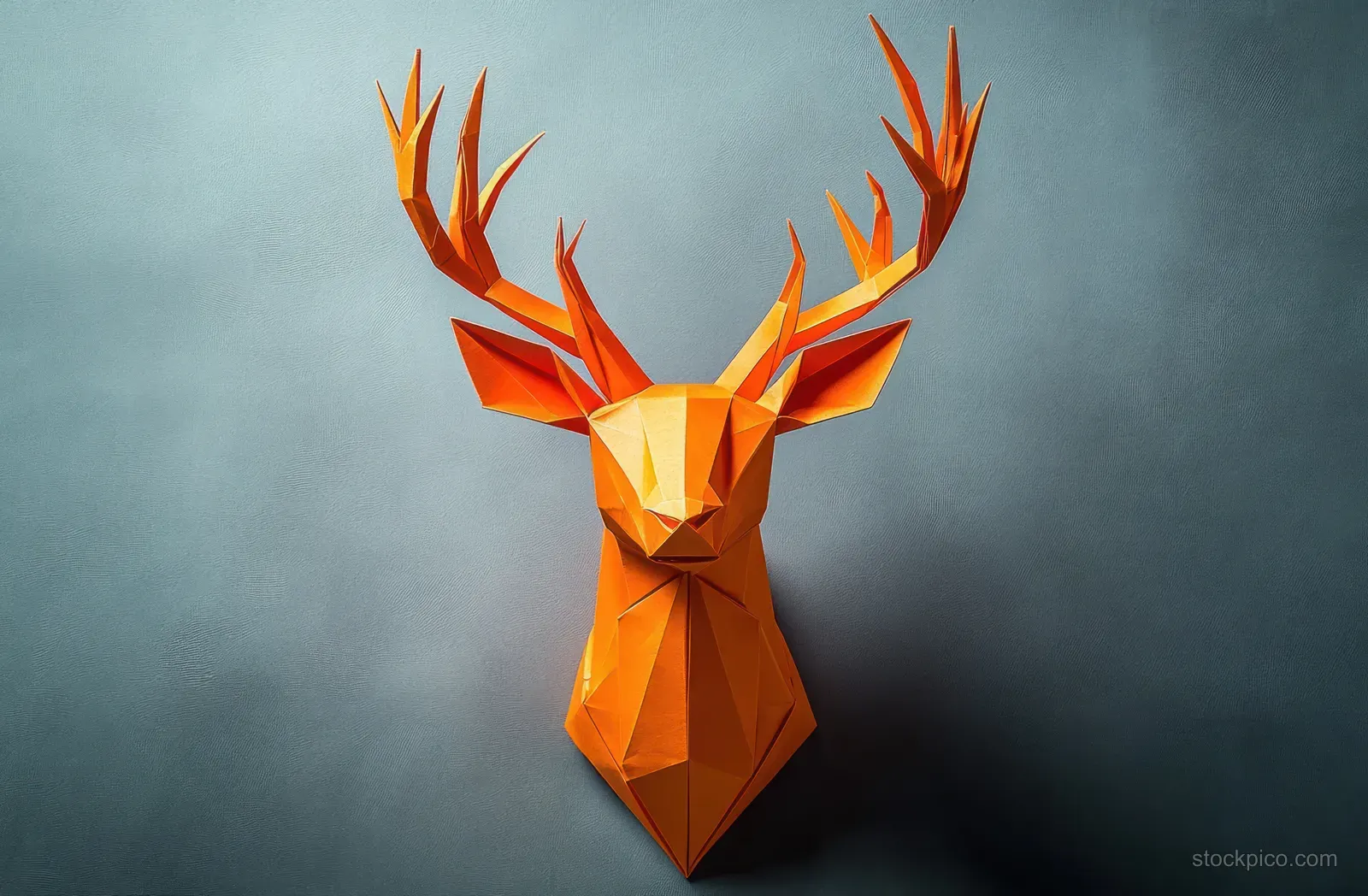 An orange geometric origami deer head sculpture mounted on a plain gray wall with visible faceted folds and shadows.