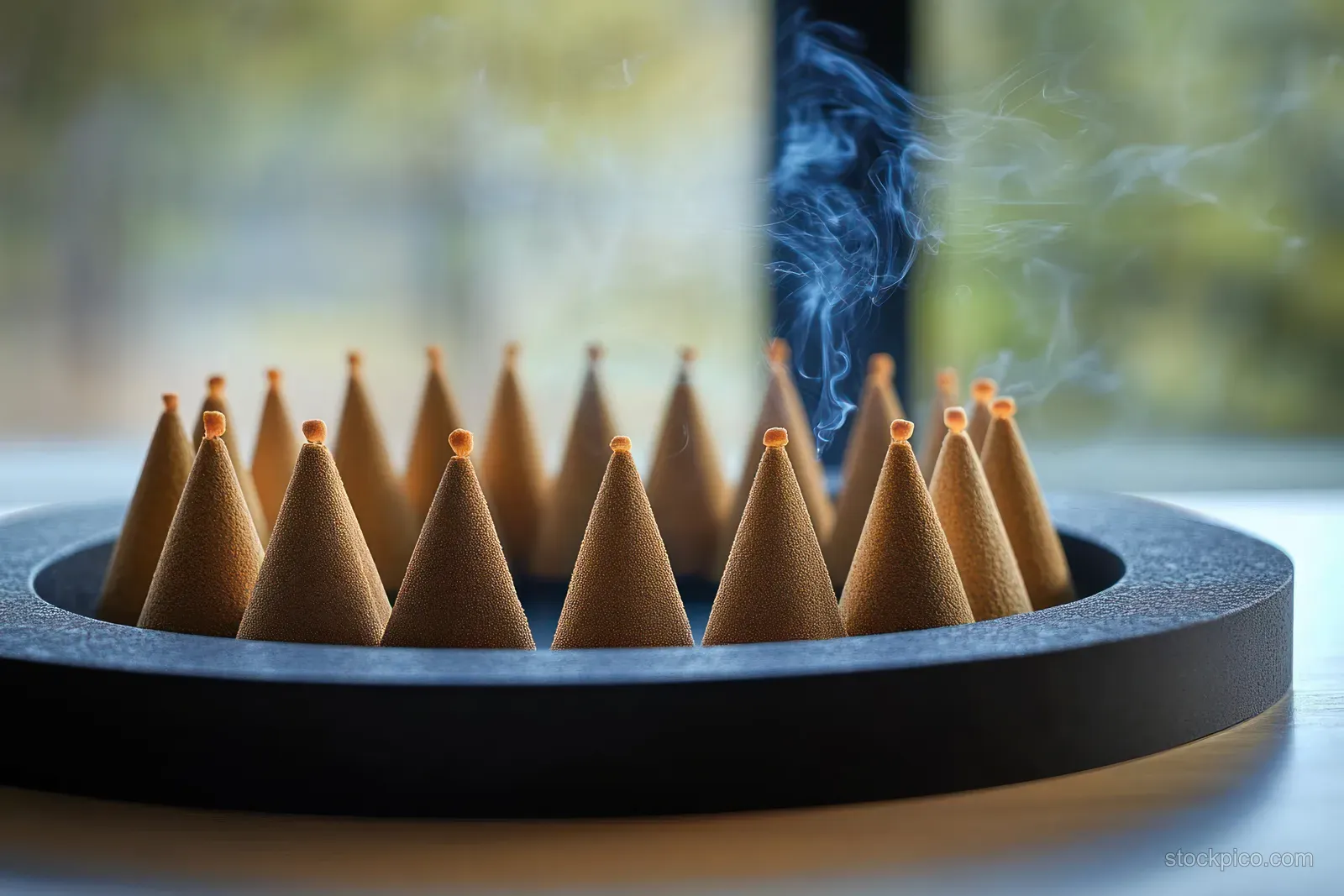 Conical moxa cones burning in a dark tray with thin smoke rising in soft daylight.
