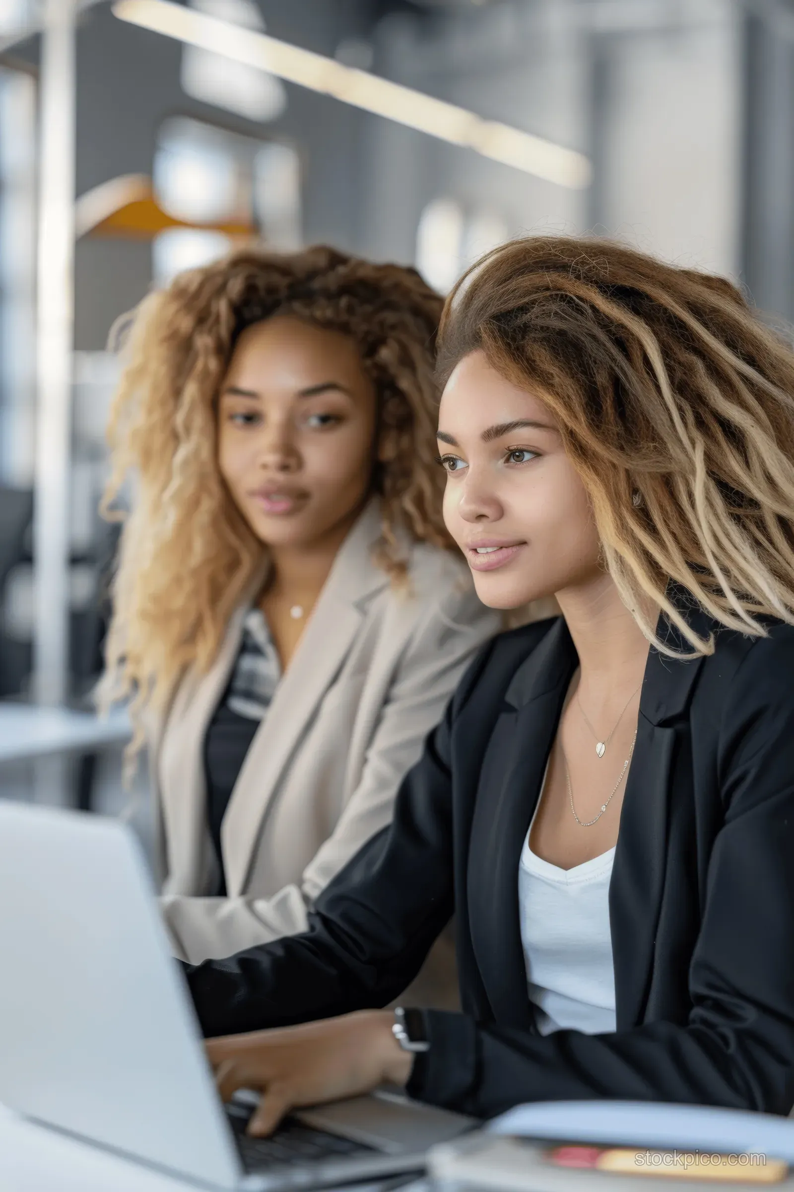 Two businesswomen collaborate at laptops in a modern office during a team meeting, with a diverse workplace visible behind them.