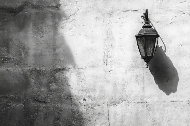 Weathered metal wall lantern mounted on a textured concrete wall with a sharp shadow cast in daylight.