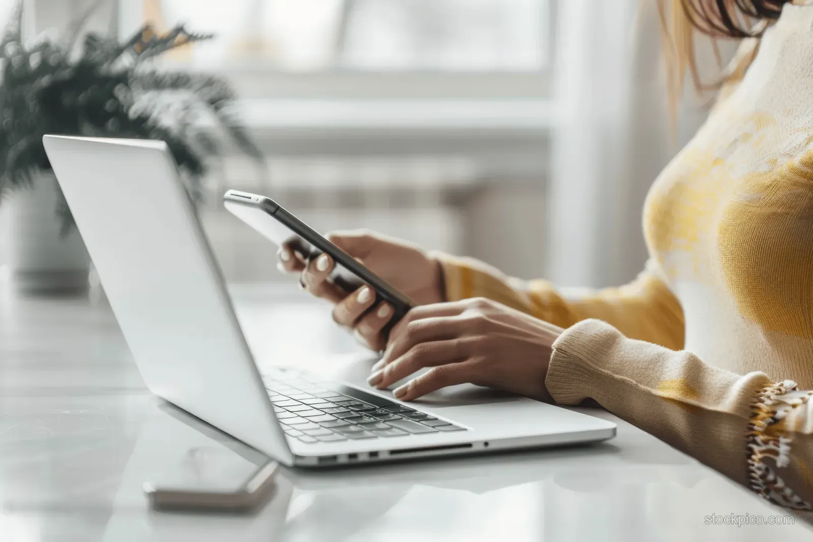 Woman types on a laptop keyboard while holding a smartphone in a minimal office with soft daylight.