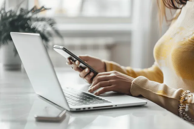Woman typing on laptop while holding smartphone in a minimal office by soft daylight