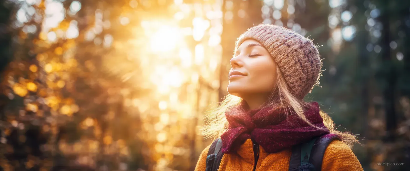 A young woman hikes along a sunlit forest trail, wearing a knit hat and scarf with a calm, reflective smile.
