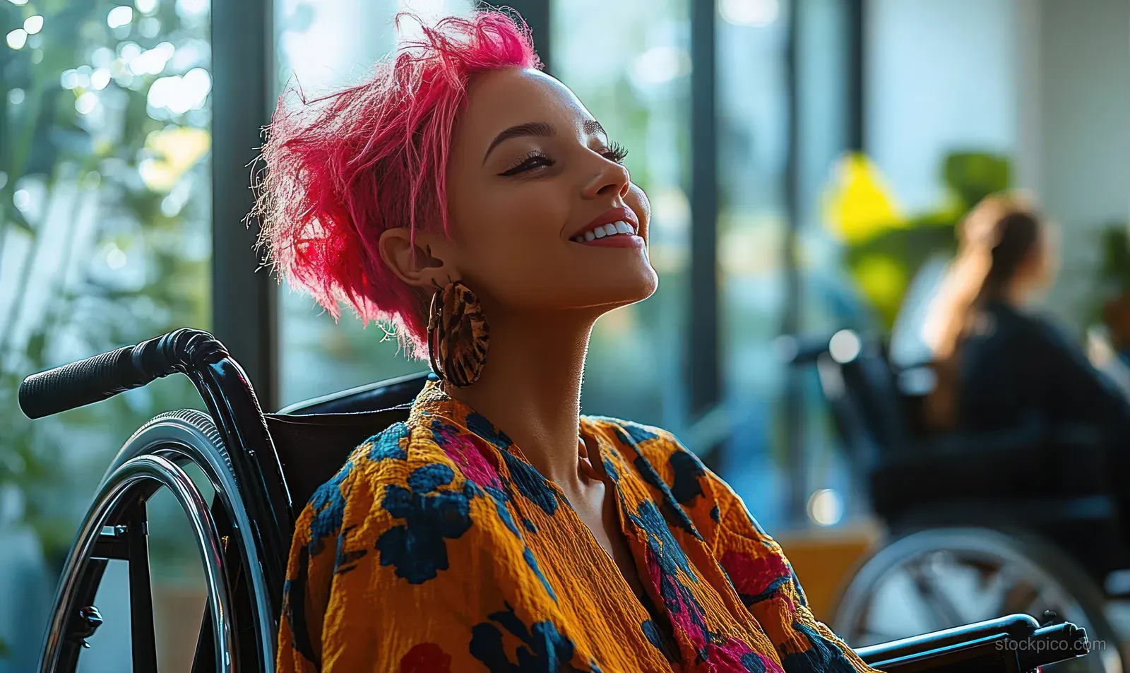 A Black woman in a wheelchair with bright pink hair smiles in a sunlit office while others work in the background.