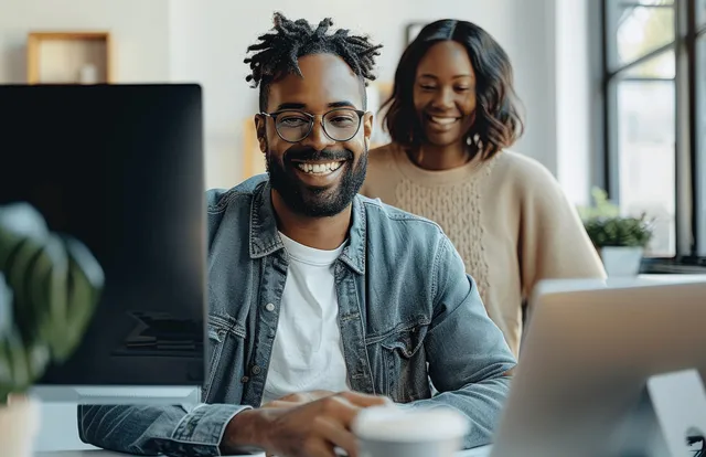 Two coworkers sit at laptops in a modern office, talking while reviewing information on screens.