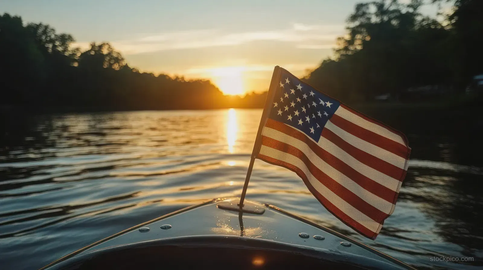 American flag waves from the front of a boat at sunset on calm lake water beside a tree-lined shore.