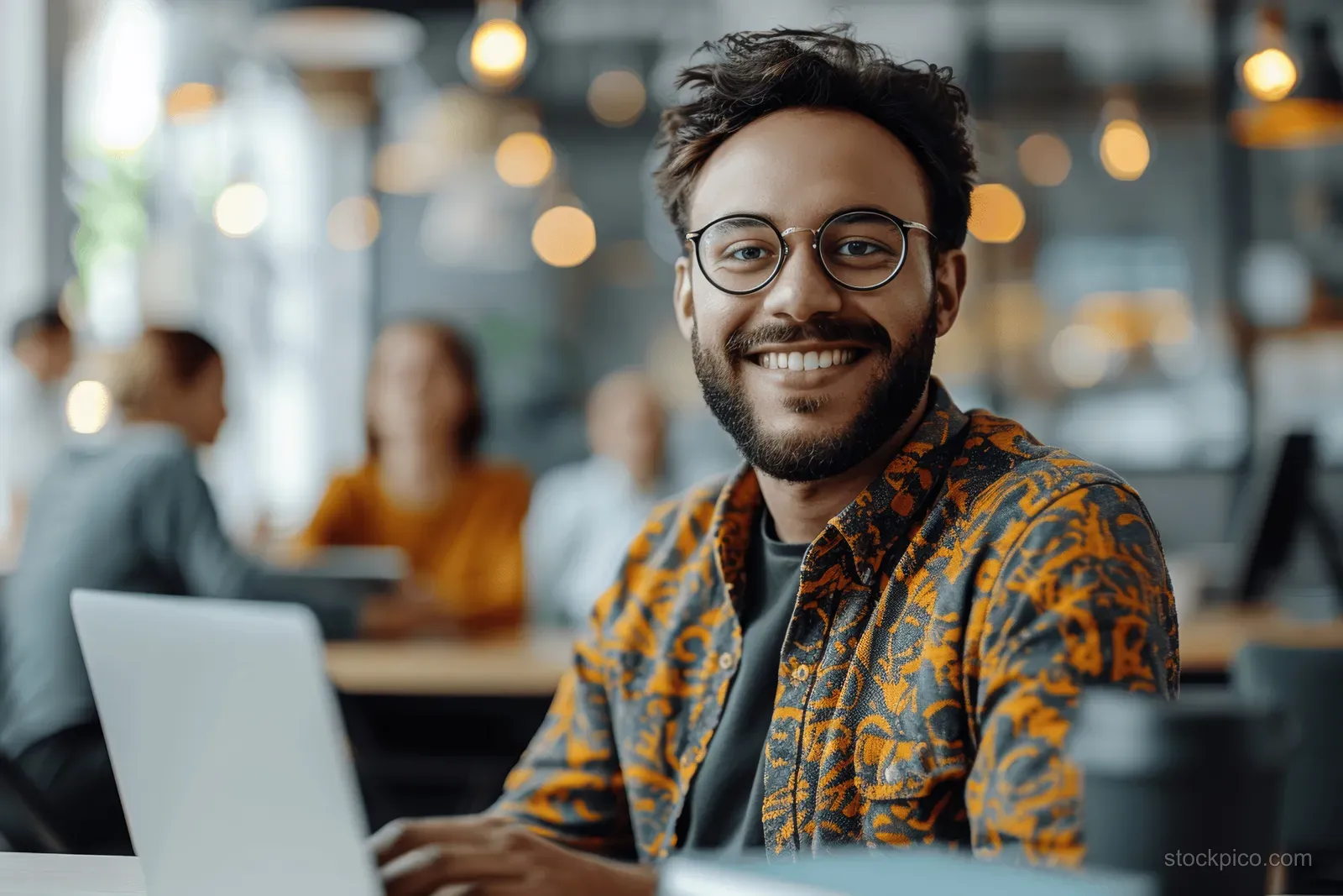 Smiling man in glasses works on a laptop at a desk while colleagues are blurred in the office background.