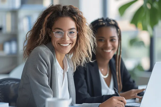 Two businesswomen collaborating on a laptop in an office, smiling during project discussion