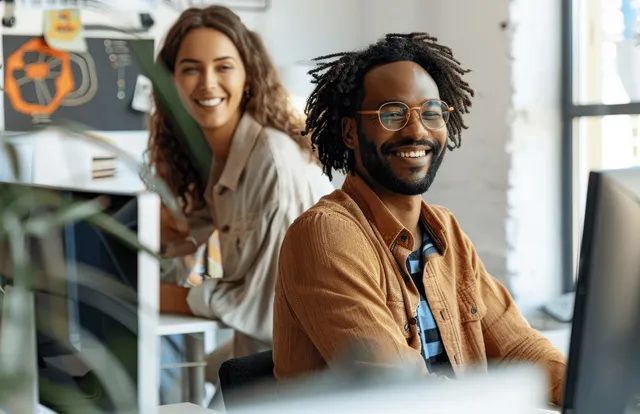 Two designers smile and review a project on computers at their desks in a bright office.