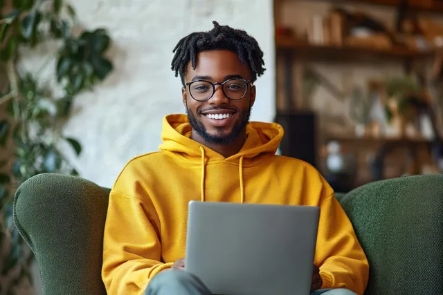 Smiling young man in a yellow hoodie using a laptop on a green armchair at home