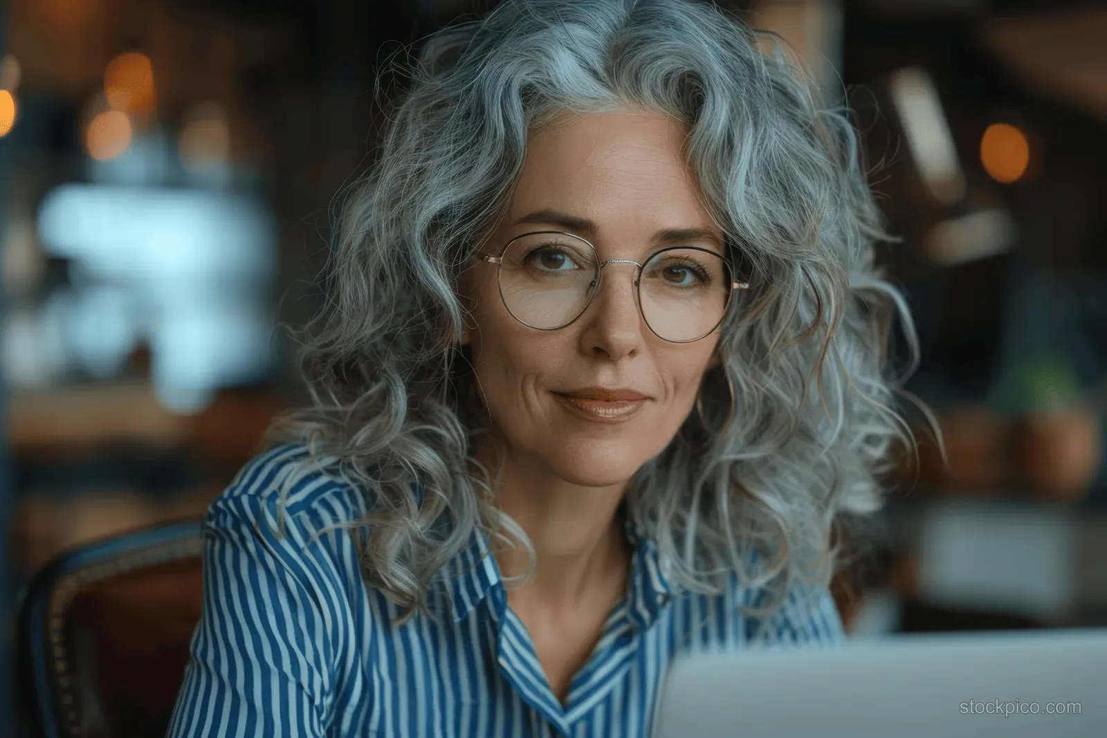 Mature businesswoman wearing glasses types on a laptop at a modern desk beside a glass wall.