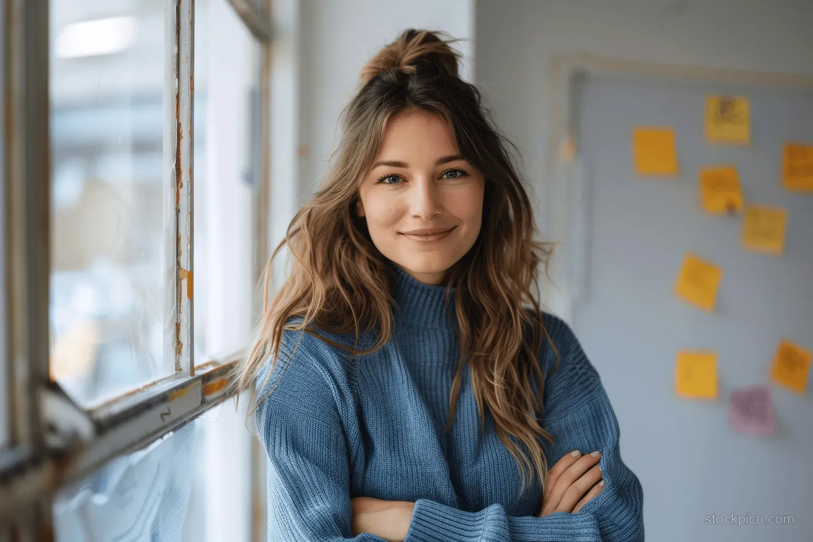 Smiling businesswoman in a blue sweater stands with arms crossed beside a window, with sticky notes on the wall behind.