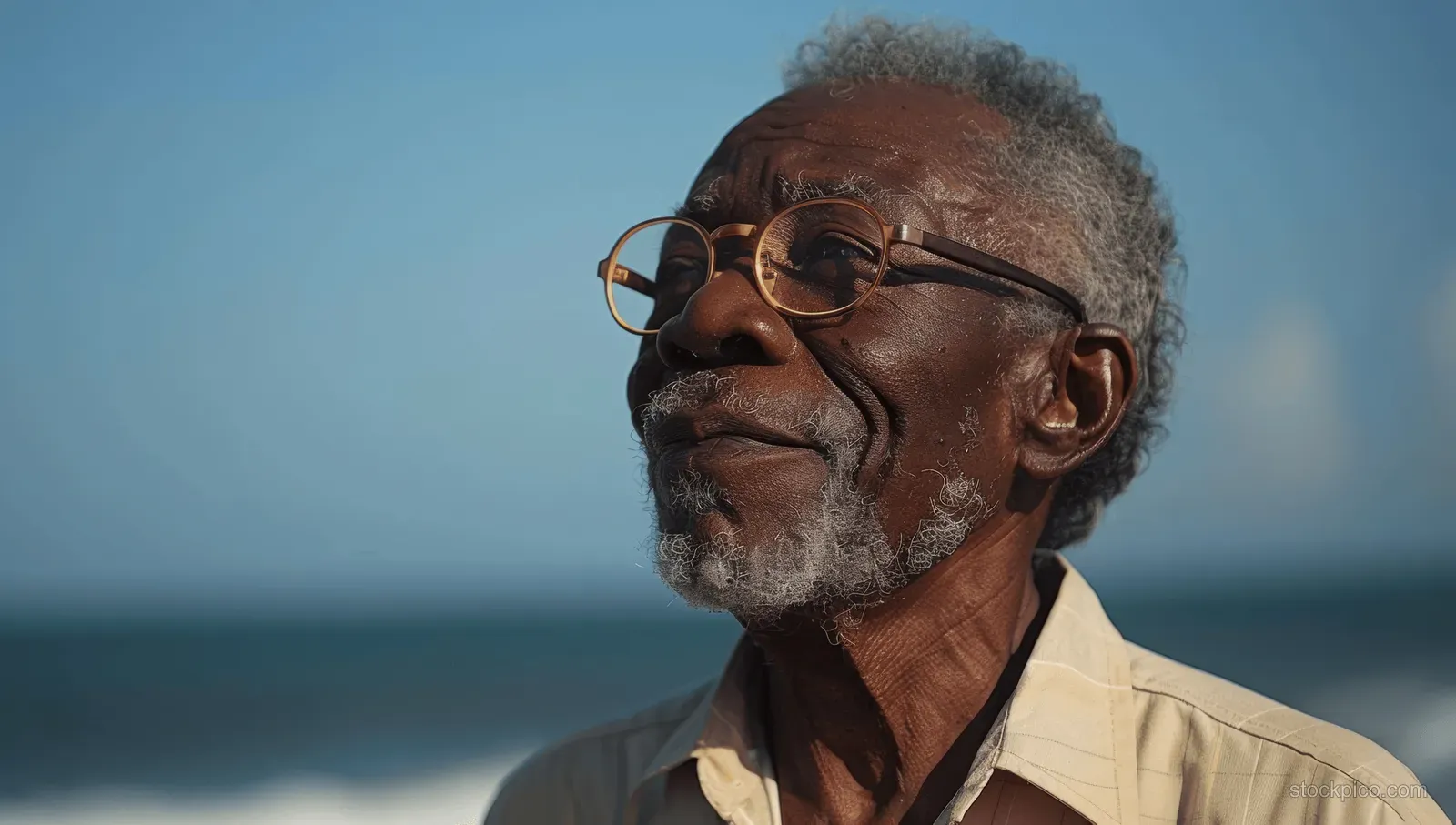 Smiling retired African American man with glasses on a sunny beach, with ocean horizon behind him.