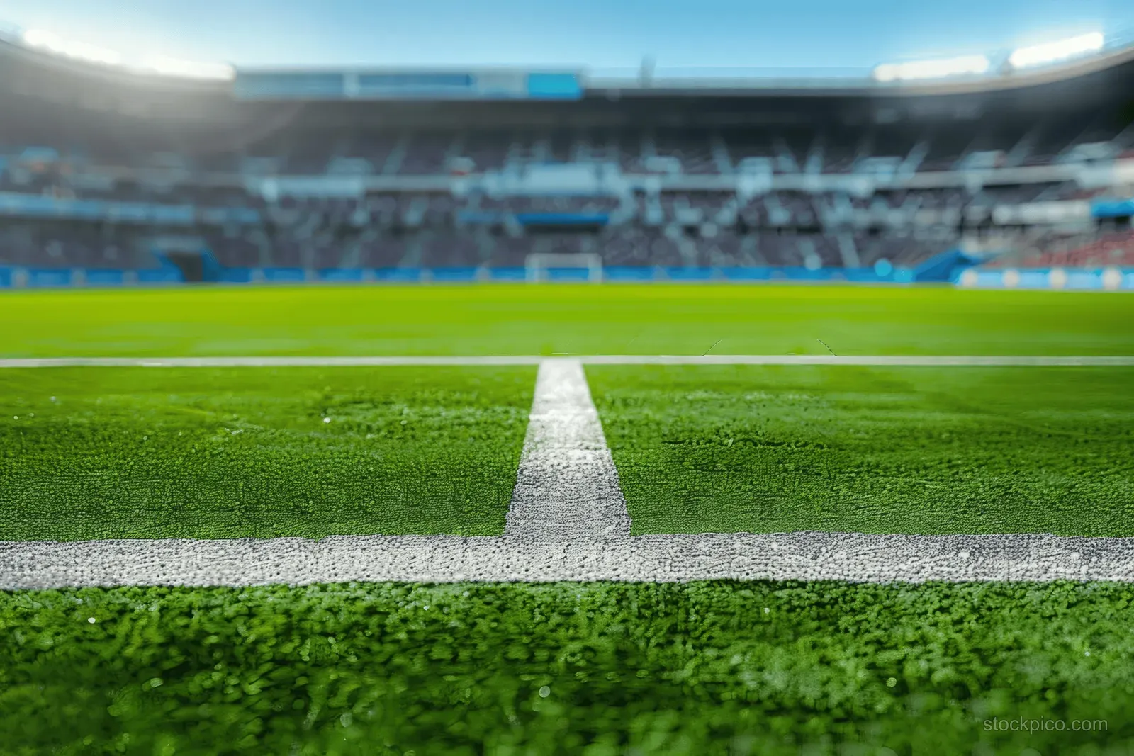 Empty soccer stadium showing bright green grass pitch with crisp white field lines and blurred stands in the background.