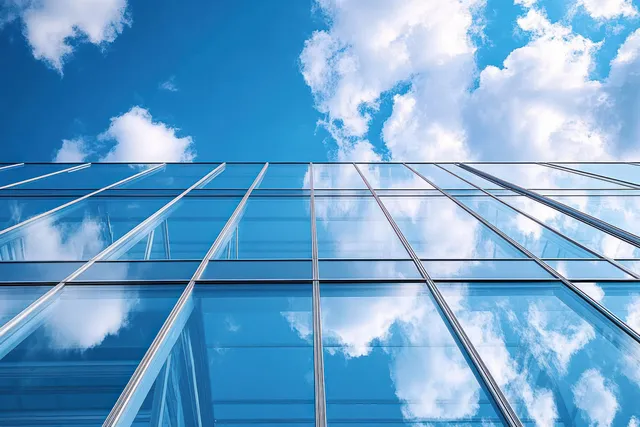Looking up at a glass building facade where blue sky and fluffy clouds reflect in the window panels.