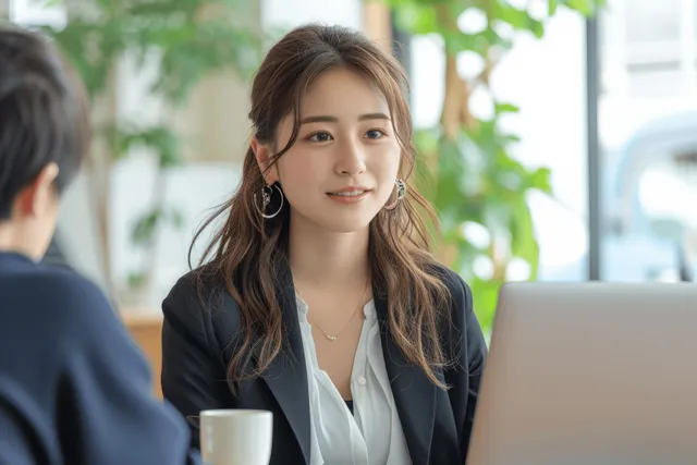 Japanese businesswoman listens in an office interview, with laptop and coffee cup on the table.