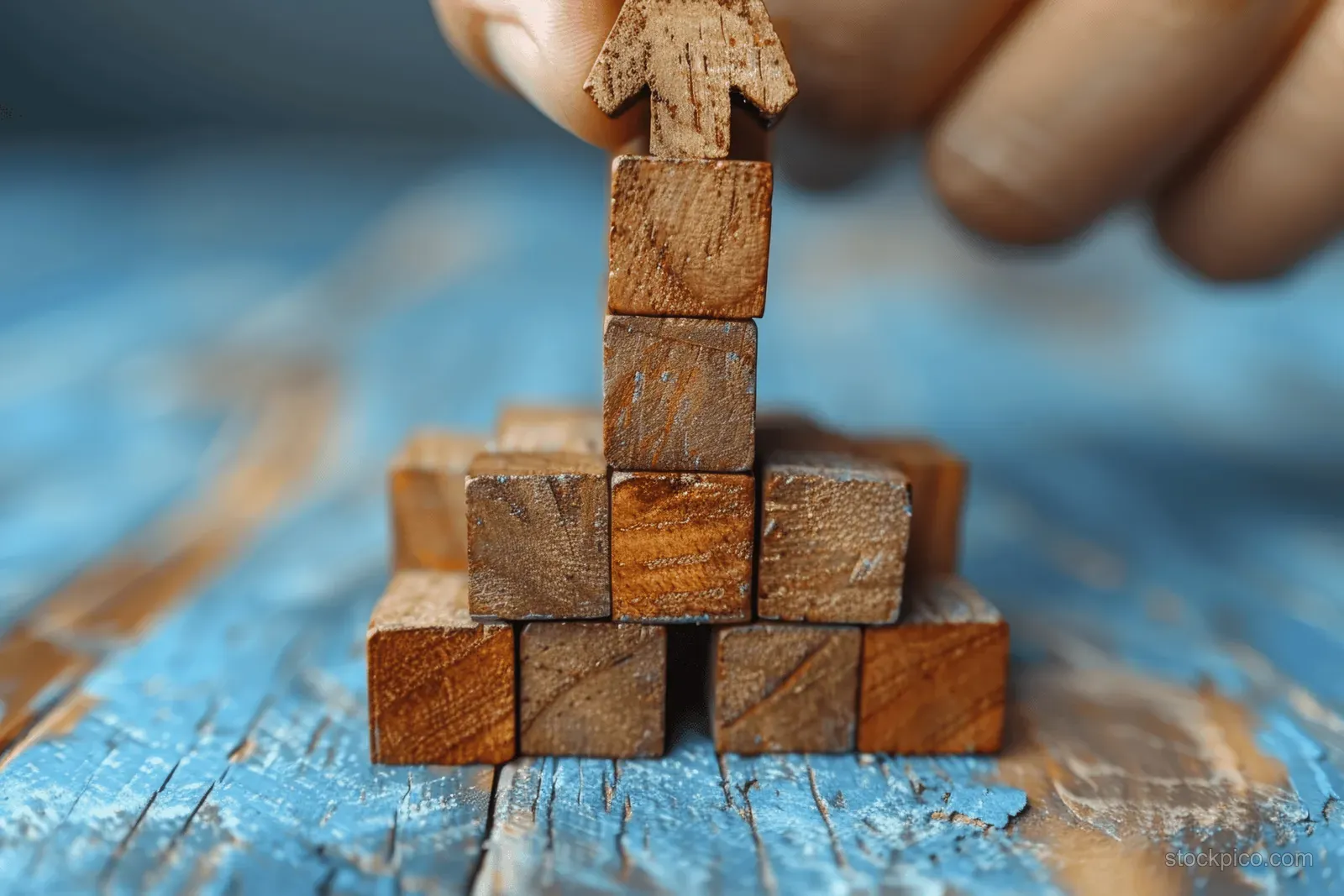 A hand stacks wooden blocks into an upward arrow on a blue wooden table.