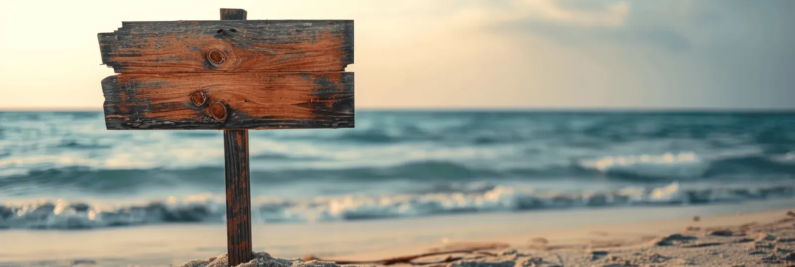 Weathered wooden beach signpost on sand with ocean waves and a blue horizon in the background.