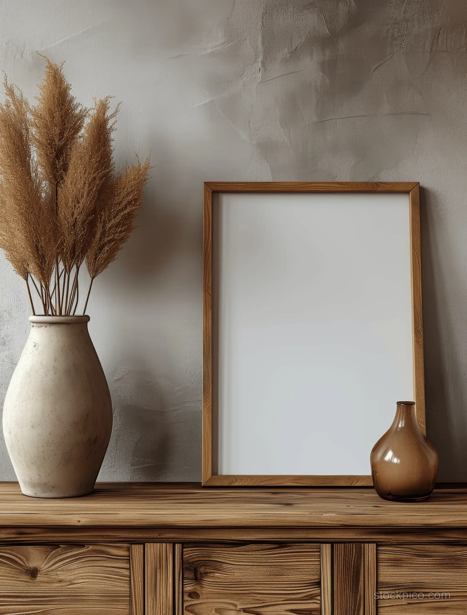 Blank wooden picture frame on a rustic console with ceramic vases and pampas grass against a beige wall.