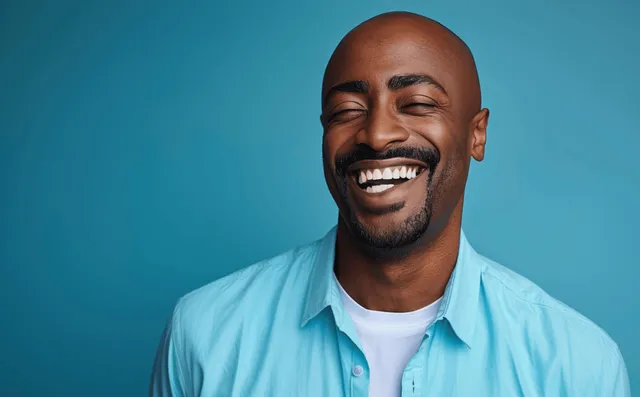 A bald man in a light blue shirt laughs against a plain cyan studio background.