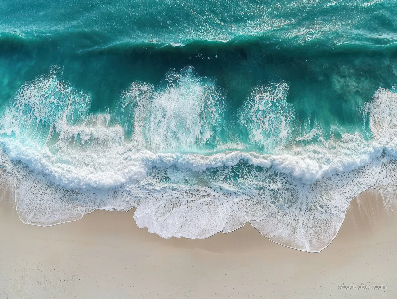 Aerial view of turquoise ocean waves breaking onto a white sandy beach with foamy surf and visible ripples.
