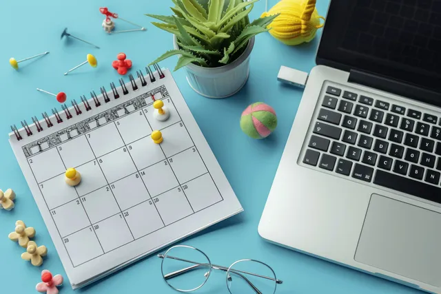 Colored pins mark dates on a calendar while an open laptop sits on a blue desk in flat lay view.