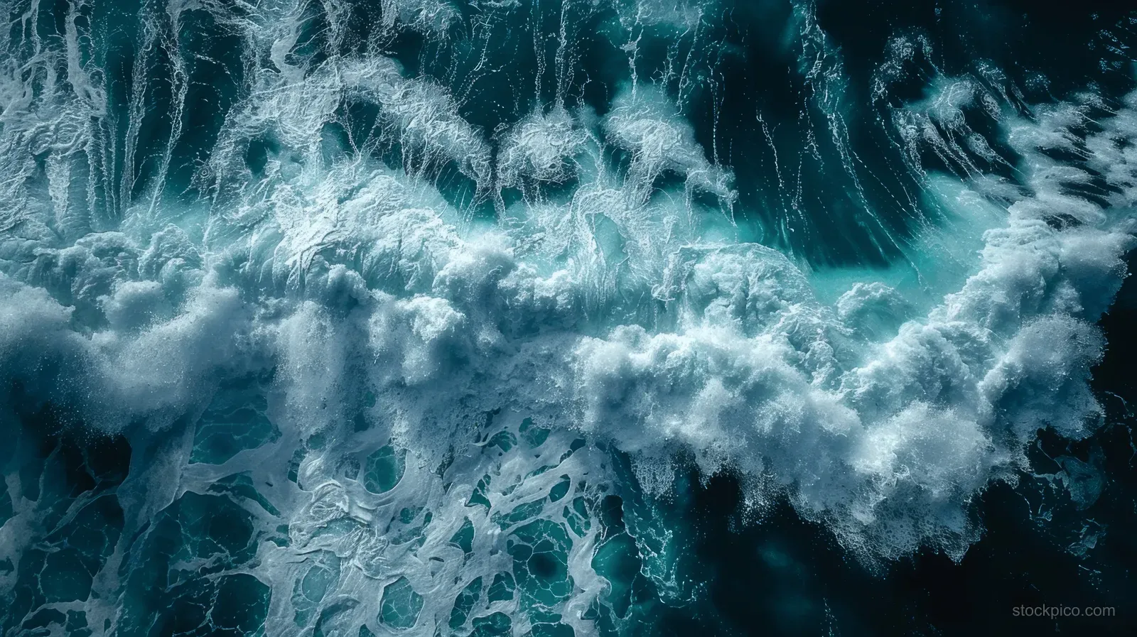 Aerial view of ocean waves with white foam forming swirling patterns over dark blue and turquoise water.