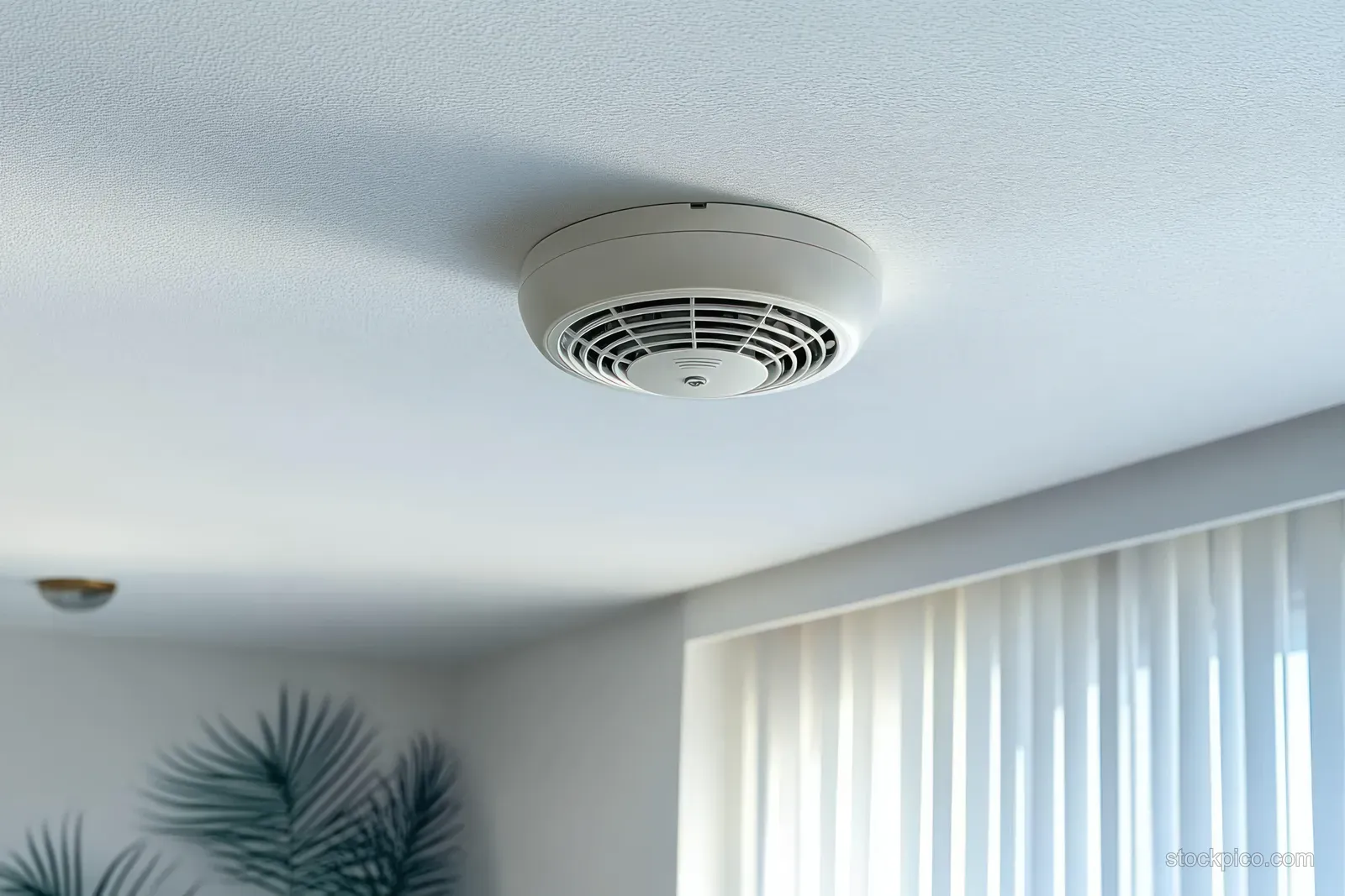 A smoke detector is mounted on a white ceiling above bright window curtains in a minimalist living room.
