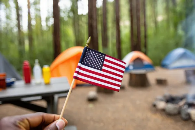 Hand holding a small American flag in front of a forest campsite with colorful tents and a picnic table.