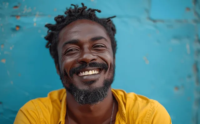 A young Black man with a short beard laughs in a minimalist studio under soft pastel lighting.