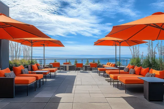 Orange lounge chairs and umbrellas line a rooftop patio overlooking a calm ocean beneath partly cloudy blue sky.