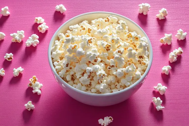 Un tazón blanco de palomitas recién hechas sobre un vibrante fondo rosa con granos esparcidos alrededor.