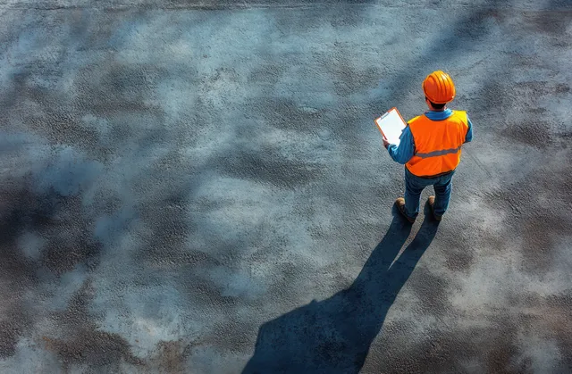 Overhead view of a construction worker in an orange safety vest walking with a clipboard across concrete with a long shadow.