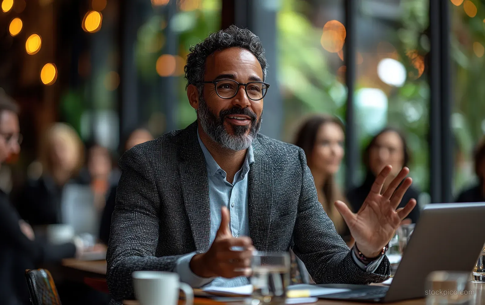 A manager gestures while leading a diverse team meeting in a modern office boardroom with laptops and coffee.