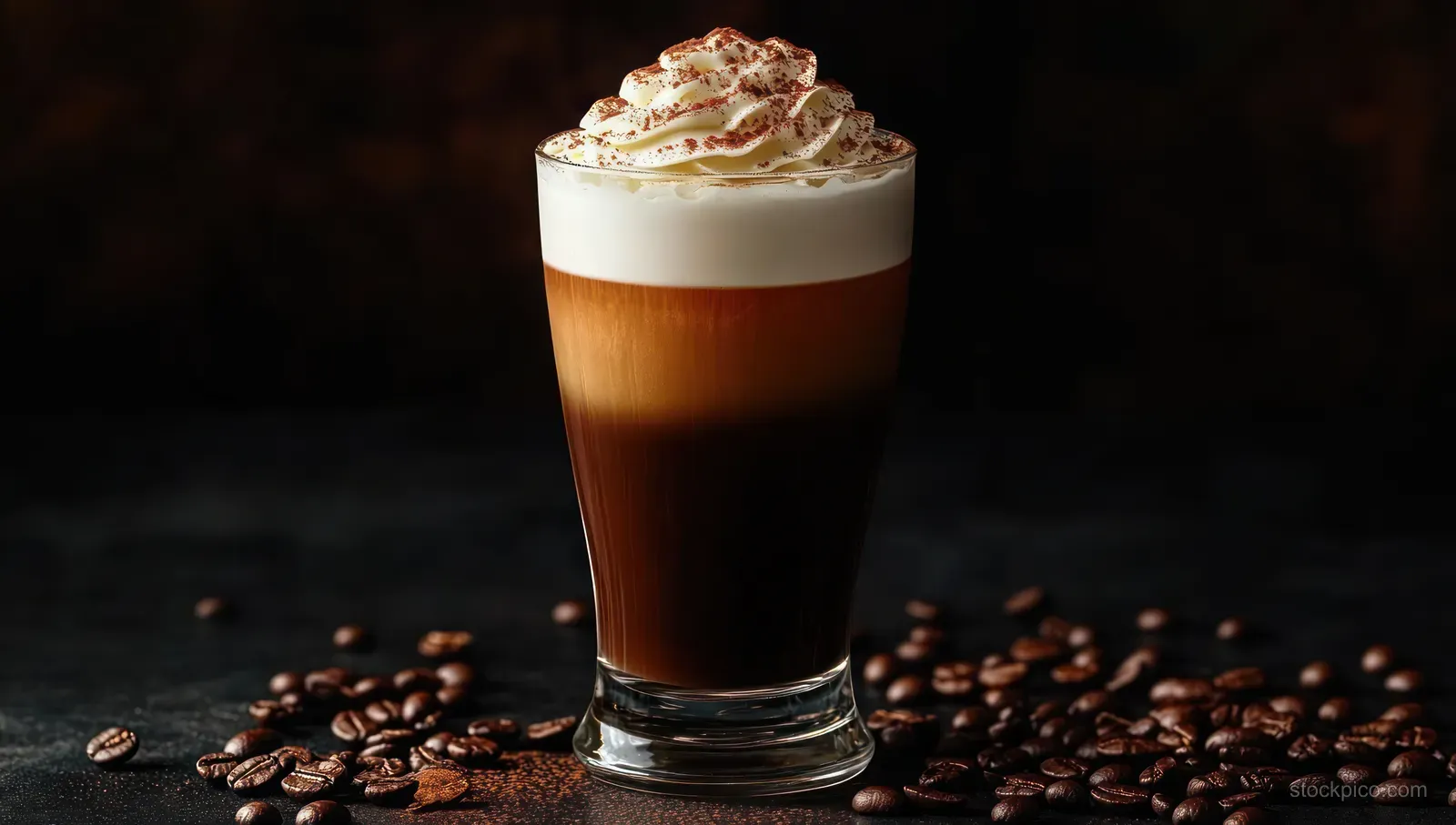 Layered iced coffee in a glass topped with whipped cream and cocoa powder, surrounded by scattered coffee beans on a dark surface.