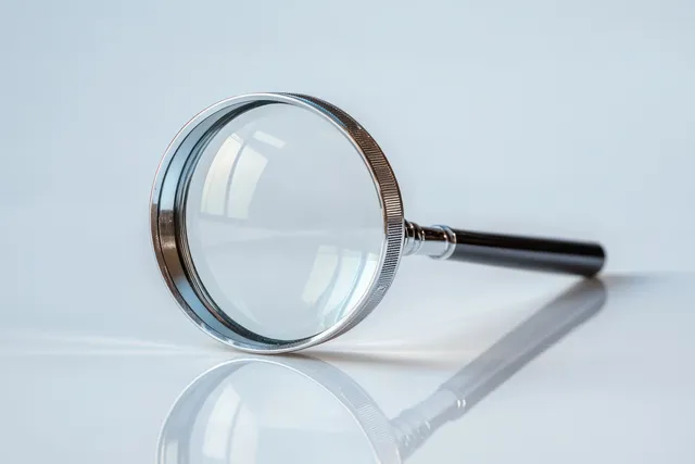 Magnifying glass with metallic rim and black handle lying on a reflective white surface, showing lens reflection and shadow.