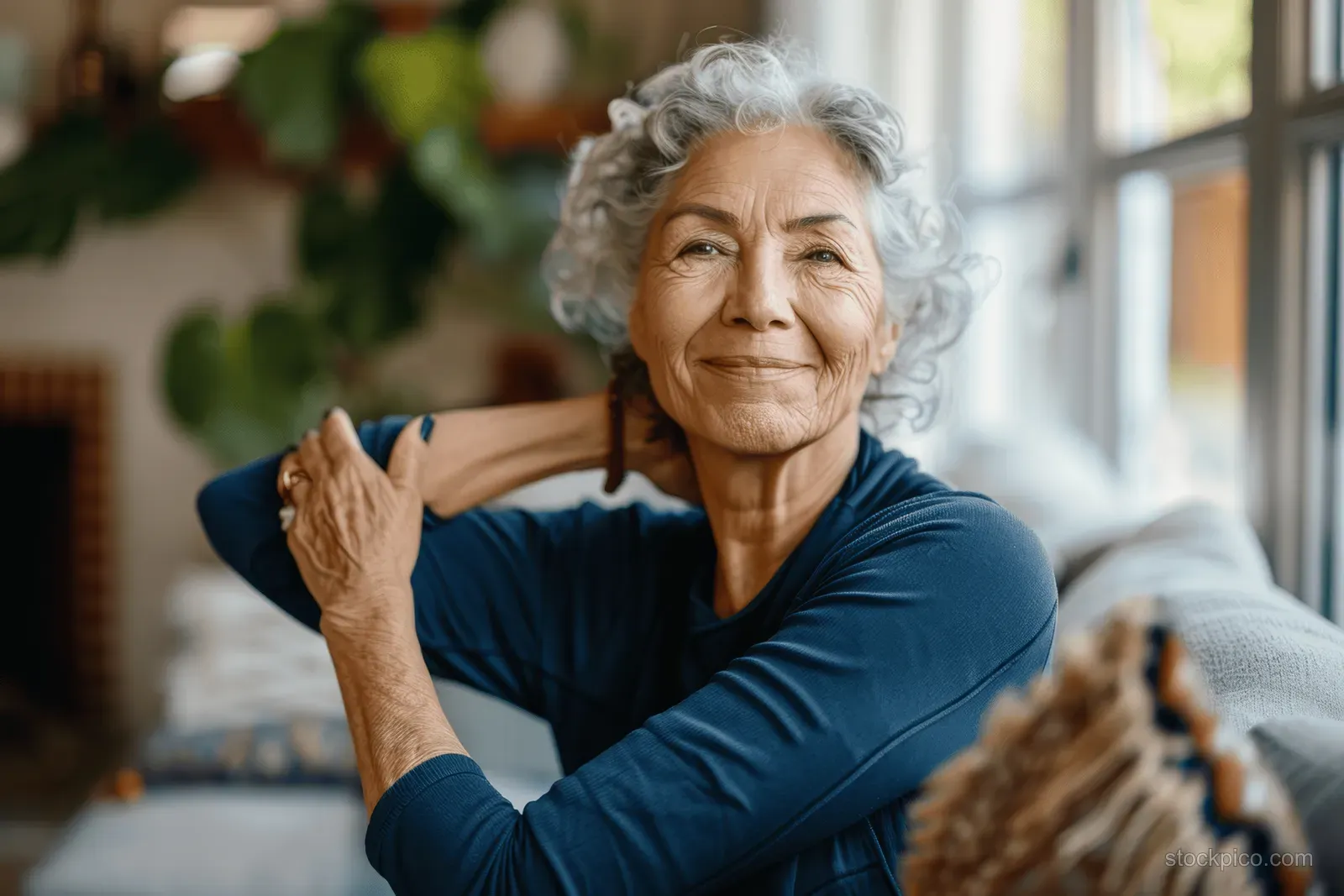 Senior woman stretching her arm across her chest while sitting on a sofa in a bright living room.