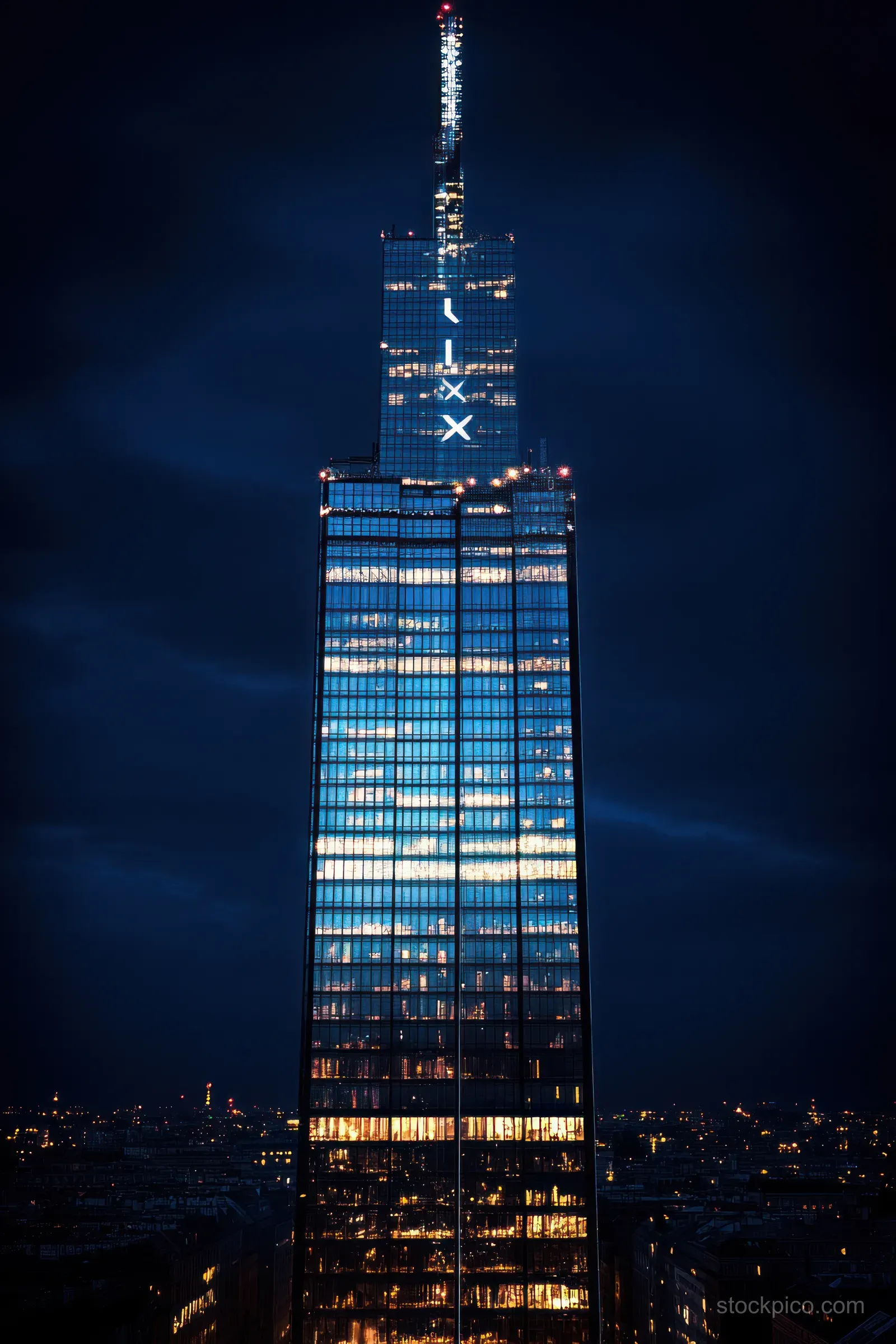 An illuminated glass skyscraper stands at night with blue city lights reflecting on its windows under cloudy skies.