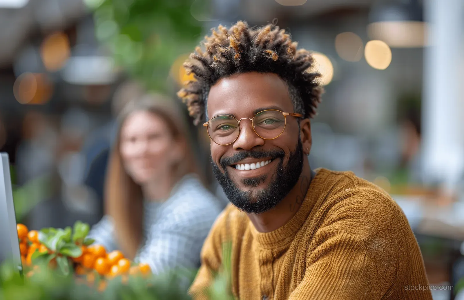 Smiling man reviews a laptop screen in a casual office with a blurred coworker in the background.