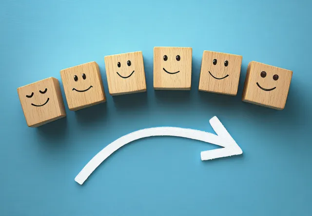 Wooden blocks with evolving smiley faces sit in sequence above a right-curving white arrow on a blue background.