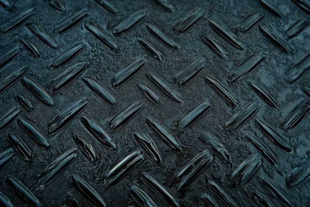 Worn dark diamond plate metal with raised tread patterns and rough reflective surface texture.