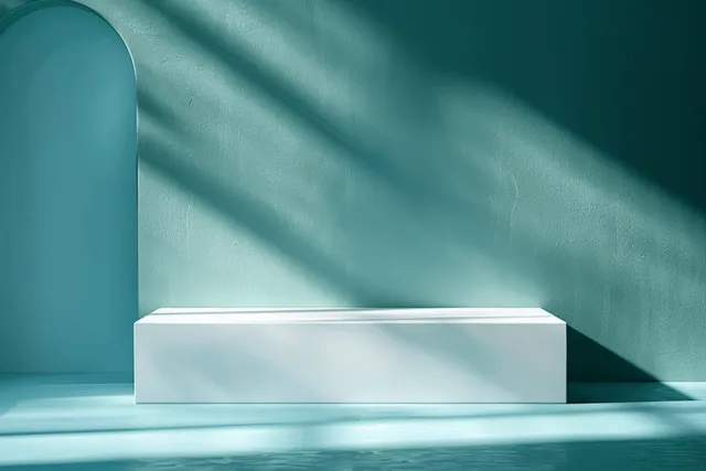 White rectangular bench sits against teal wall in a minimalist interior with dramatic sunlight shadows.