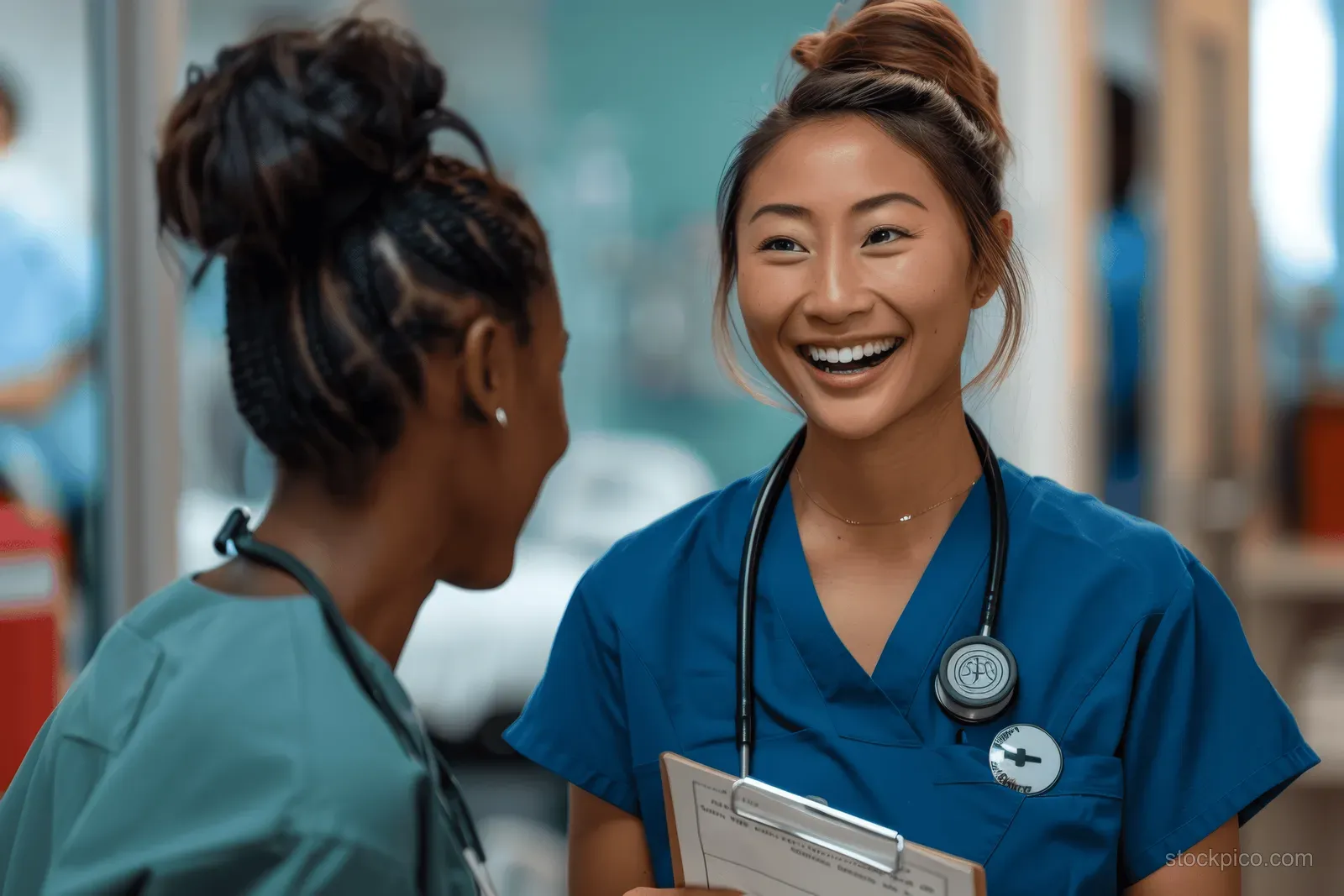 Smiling female medical professional in blue scrubs chats with a colleague in a hospital hallway while holding a clipboard.