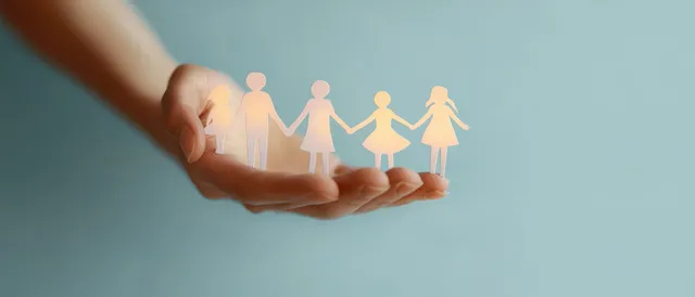 A caregiver’s hand holds small diverse family and children icons against a soft blue background.