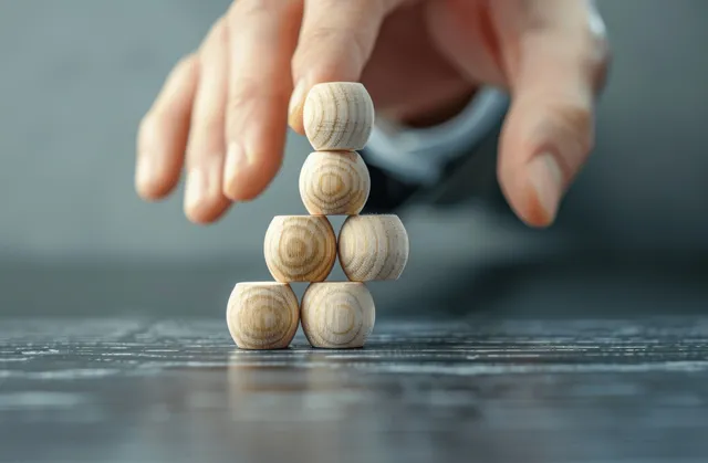 A hand places a wooden block on a growing stack with target and arrow elements on a gray background.