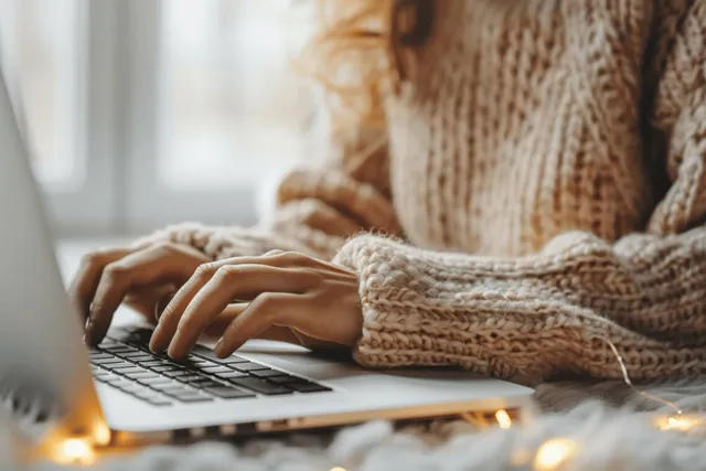 Close-up of a woman’s hands typing on a laptop keyboard at a home desk with warm, softly blurred background.