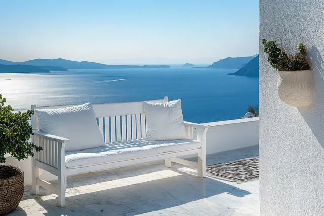 White bench with cushions on a seaside terrace overlooking calm blue sea and distant mountains.