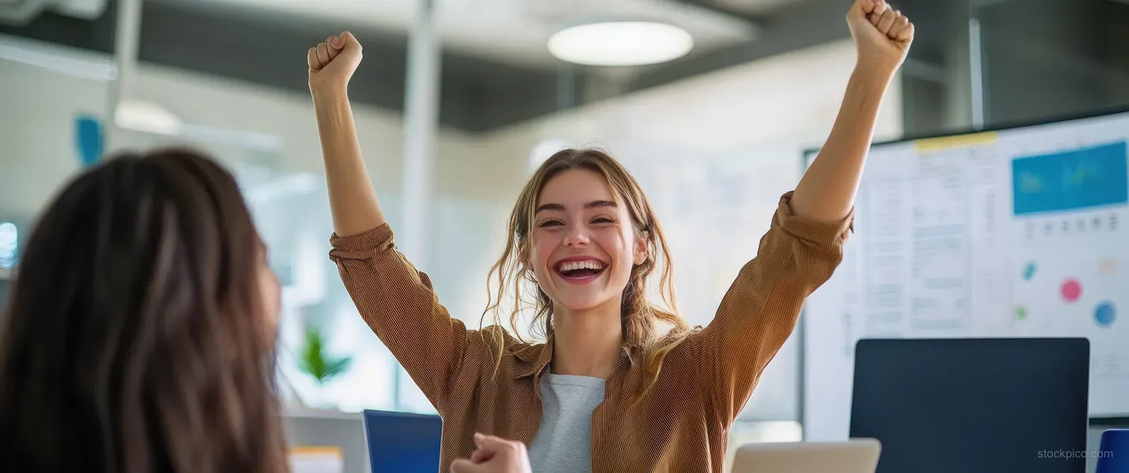 A young woman celebrates with raised arms and a joyful smile in a bright modern office.