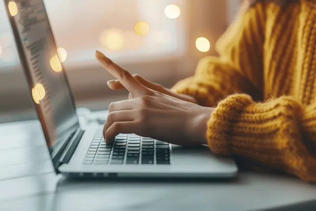 A woman types on a laptop keyboard at her desk with warm office lights blurred in the background.