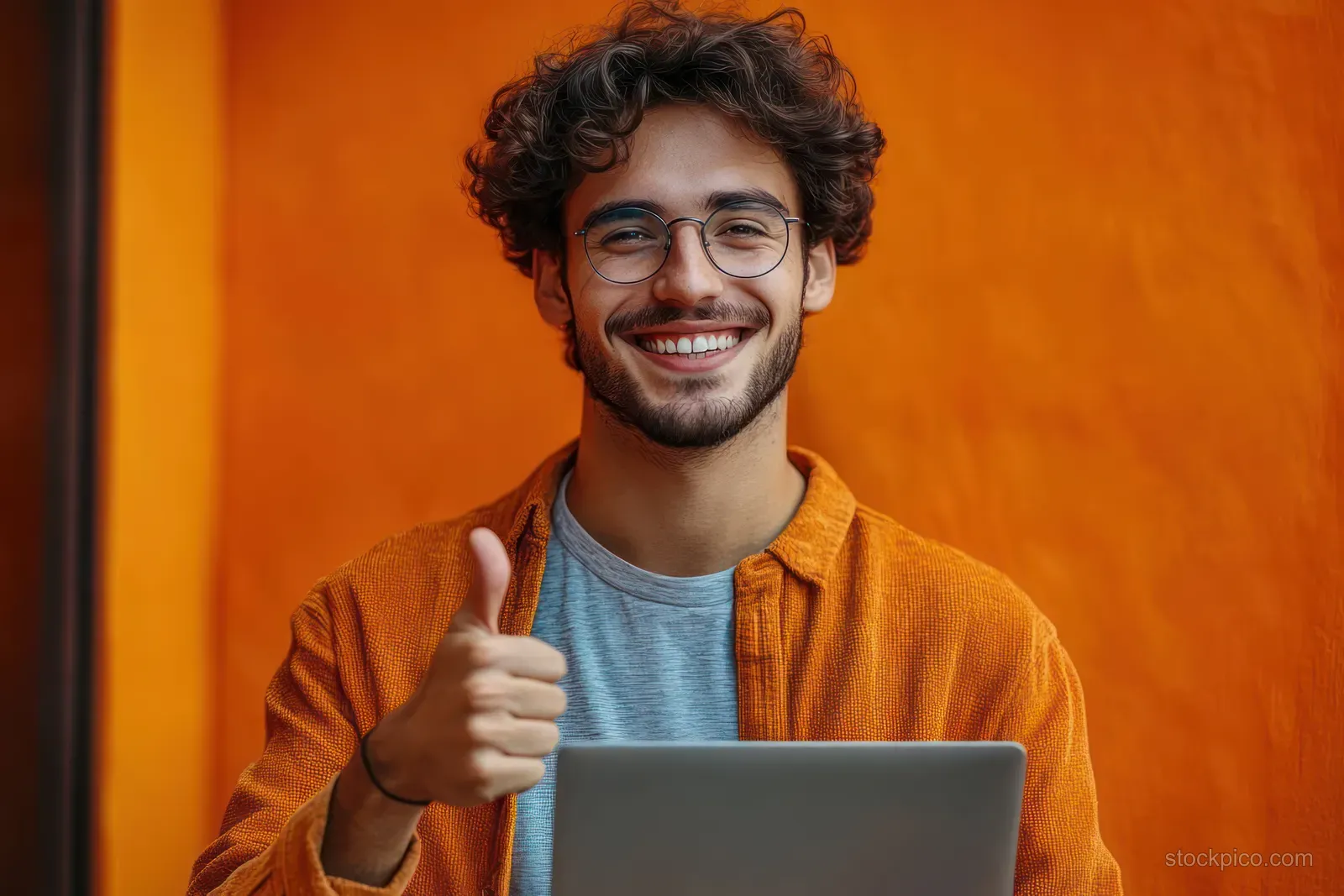 Curly-haired man wearing glasses gives a thumbs up while using a laptop against a bright orange background.